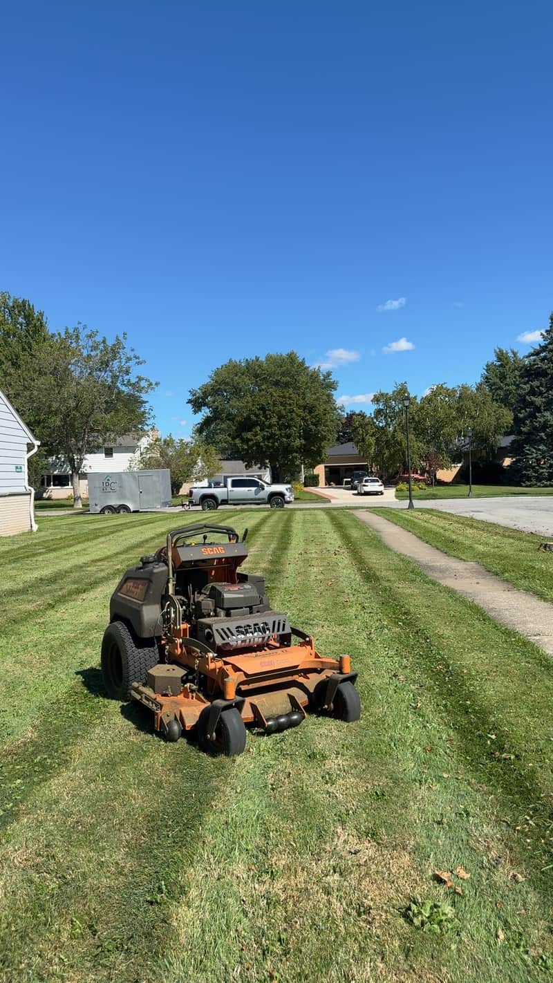An orange stand-on mower cutting stripes across a residential front lawn on a quiet street
