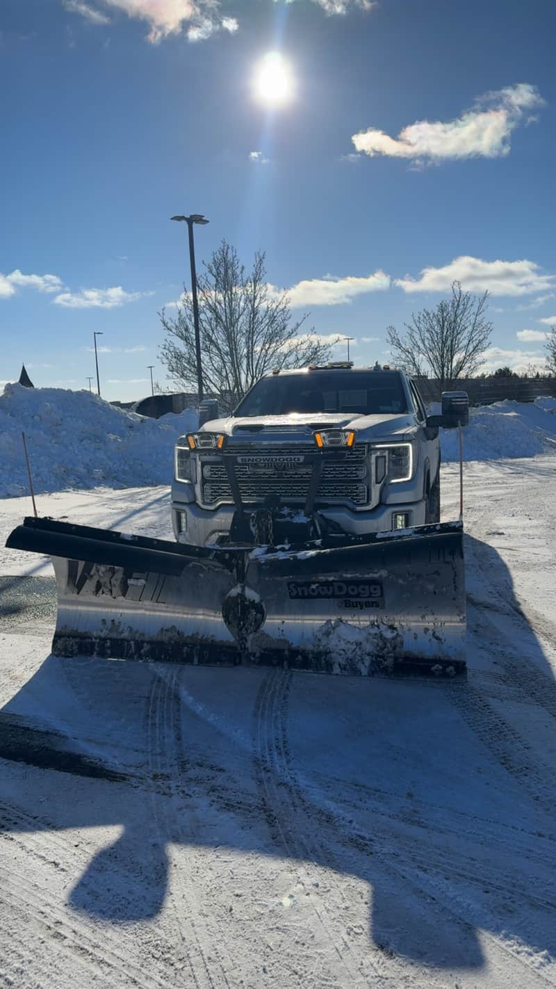 A pickup truck fitted with a snow plow blade on a freshly cleared lot beside a bank of pushed snow
